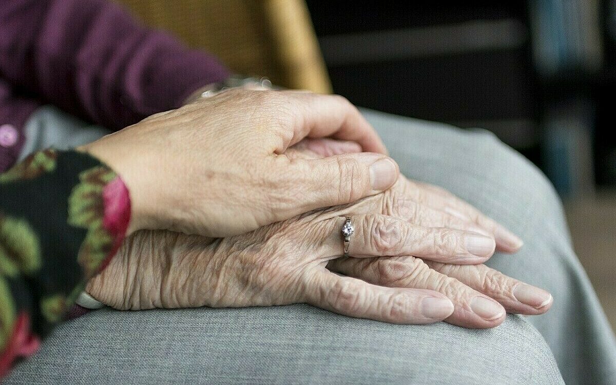 Eine Hand liegt auf zwei Händen einer Seniorin auf dem Schoß. Eine Hand liegt auf zwei Händen einer Seniorin auf dem Schoß.